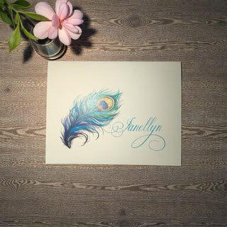 Card with a peacock feather design and a personalized name laying on a wooden surface with a pink flower.